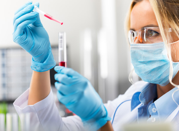 A health practitioner in a lab, with a lab mask covering her face. Focusing on a test tube and a sample in her hands.